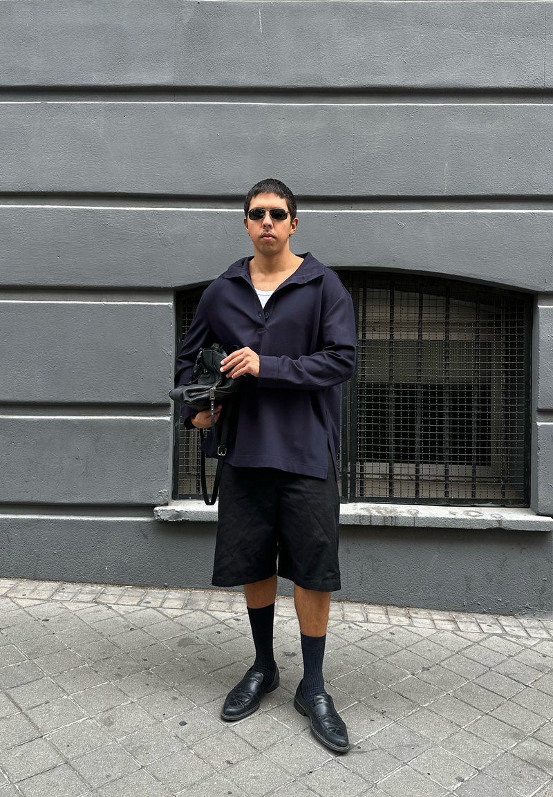 Pulôver oversized azul-marinho com gola grande, calções pretos, meias pretas e sapatos; segurando uma pequena bolsa preta contra uma parede cinza.