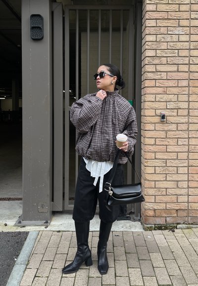 Mujer con gafas de sol que lleva una chaqueta de cuadros, pantalones cortos negros hasta la rodilla, botas negras, sosteniendo una taza de café y un bolso negro junto a una pared de ladrillos.