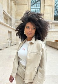 Veste bomber beige sur un T-shirt blanc avec un détail en nœud rose, assortie à un pantalon beige. Cheveux noirs bouclés, cadre intérieur.