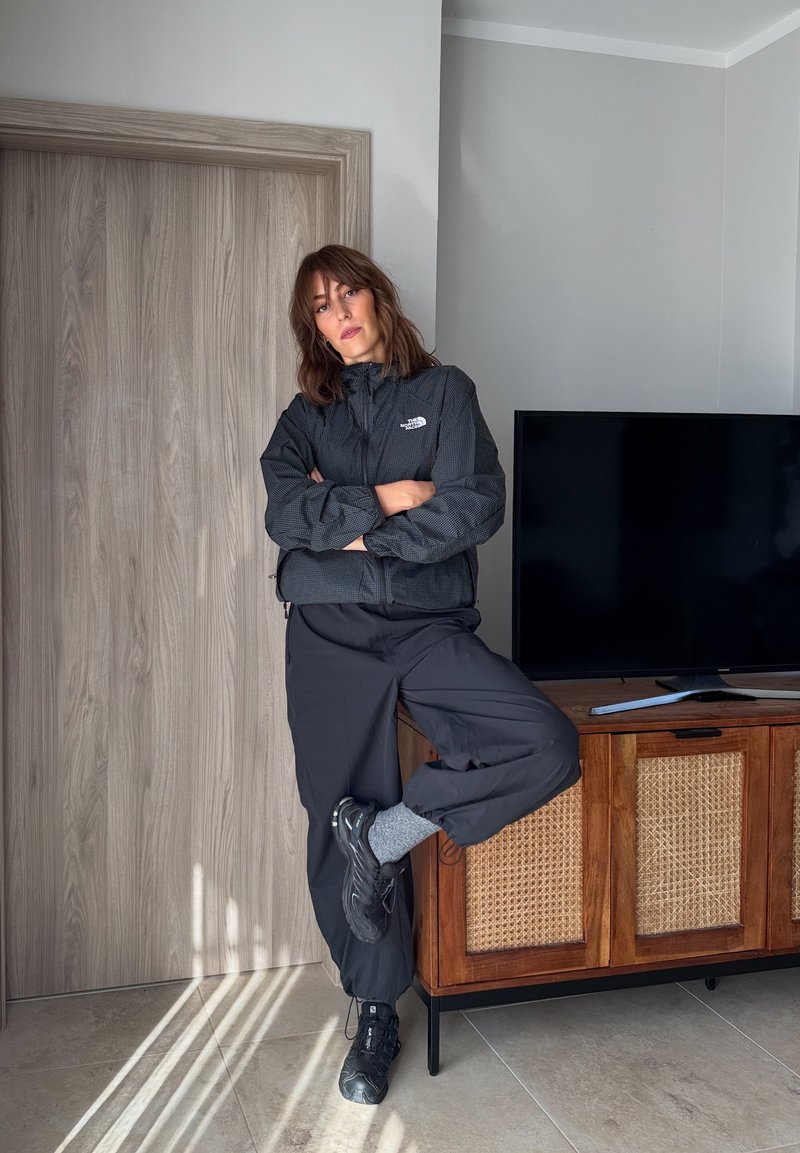 Une femme vêtue de vêtements d’extérieur foncés s’appuie contre un meuble TV en bois, les bras croisés et un pied levé, dans un salon lumineux.
