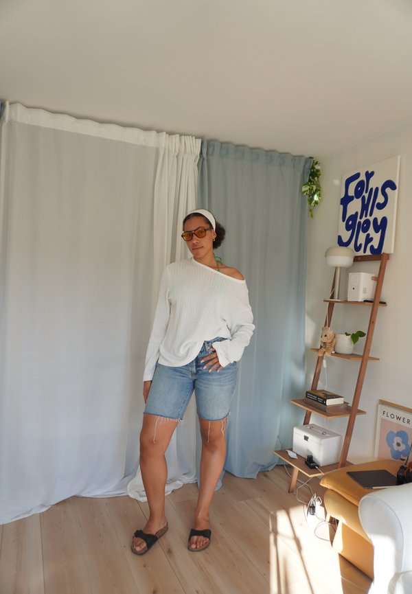 Bela oversize pulover, denim kratke hlače, rjave natikače in okrogla rumena sončna očala. Nevtralna kulisa z modrimi in belimi zavesami.