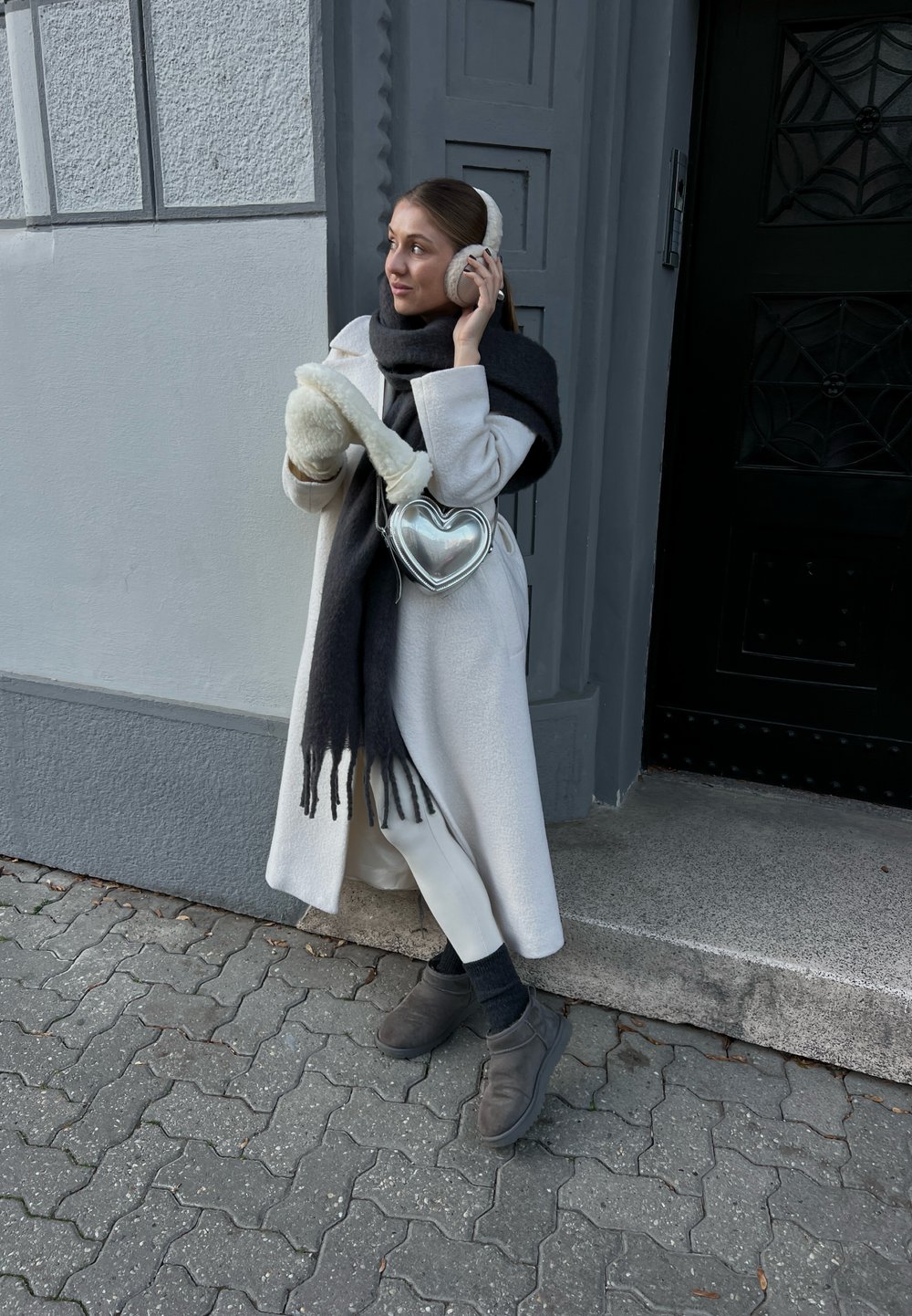 Λευκό παλτό, μαύρο κασκόλ με φούντες, ασημένια τσάντα σε σχήμα καρδιάς, κρεμ χειροπετσέτες αυτιών, κρεμ γάντια, γκρι παπούτσια, στέκεται μπροστά σε έναν γκρι τοίχο.