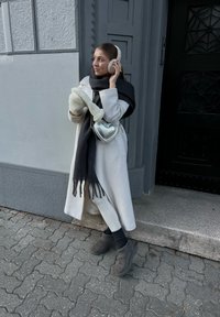 Λευκό παλτό, μαύρο κασκόλ με φούντες, ασημένια τσάντα σε σχήμα καρδιάς, κρεμ χειροπετσέτες αυτιών, κρεμ γάντια, γκρι παπούτσια, στέκεται μπροστά σε έναν γκρι τοίχο.