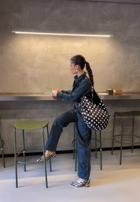 Γυναίκα με πλεγμένα μαλλιά, που φοράει σκοτεινό τζιν, τακούνια με σχέδιο ζέβρας και τσάντα με πουά, στέκεται δίπλα στον πάγκο κρατώντας έ να λευκό φλιτζάνι σε ένα μινιμαλιστικό καφέ.