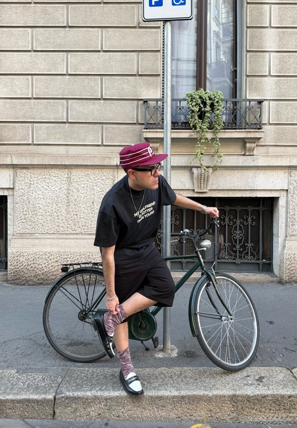 Hombre con gorra granate y ropa negra ajustándose el calcetín mientras se apoya en una bicicleta verde junto a un poste con una señal de aparcamiento para discapacitados.