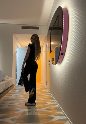 Conjunto negro ajustado, suelo con un patrón en naranja y gris, televisor curvado montado en la pared con un acento rosa, cabello largo y bolso negro.