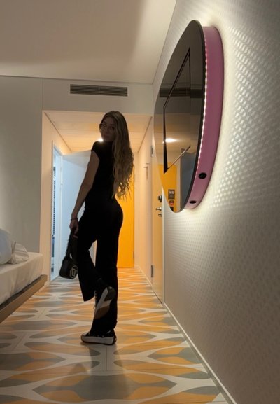 Conjunto negro ajustado, suelo con un patrón en naranja y gris, televisor curvado montado en la pared con un acento rosa, cabello largo y bolso negro.