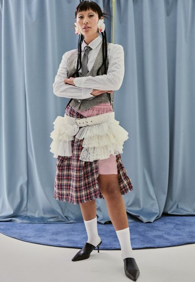 Modelo con un chaleco gris sobre una camisa a rayas, llevando una falda de tela mixta con detalles de cuadros y encaje, junto con tacones negros de punta y calcetines blancos.