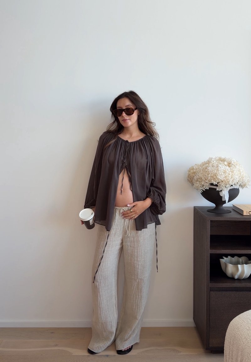 Femme portant des lunettes de soleil, une blouse marron ample et un pantalon beige, tenant un café, touchant son ventre découvert, debout près d'une table avec des fleurs blanches.