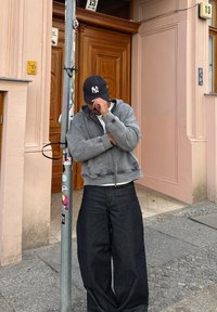 Grey zip-up hoodie with a textured finish, worn over a white shirt. Paired with loose-fitting dark denim jeans. Black baseball cap.