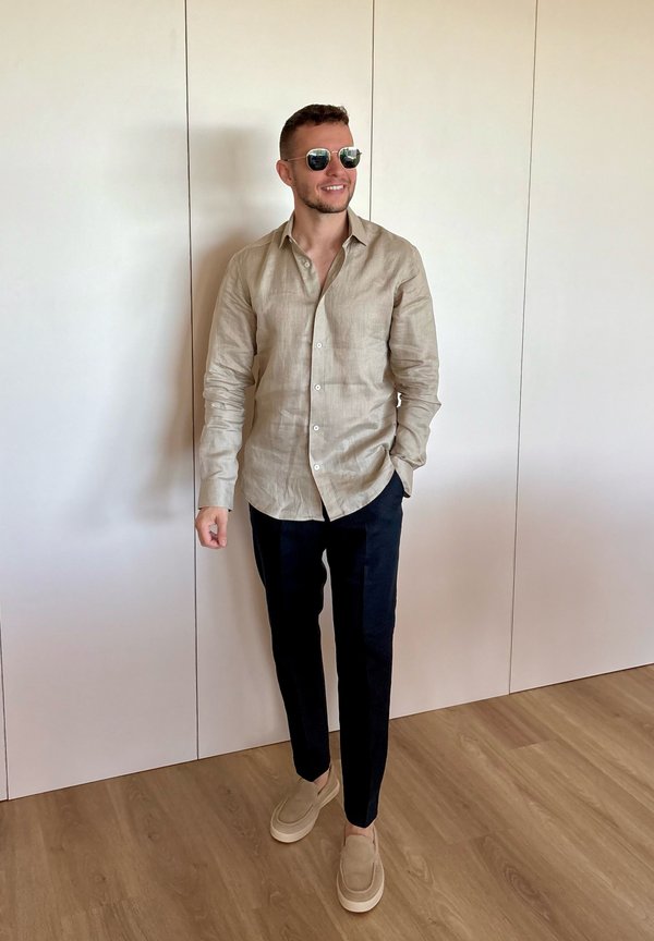 Homme portant une chemise beige boutonnée, un pantalon noir, des chaussures beiges et des lunettes de soleil, debout sur un sol en bois devant un mur blanc.