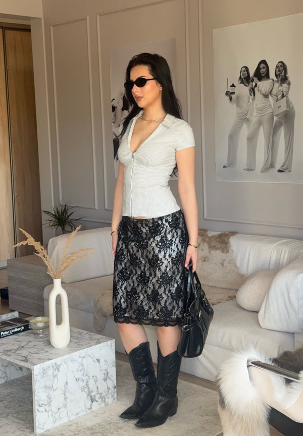 Vrouw met zonnebril, wit getailleerd topje, zwarte kanten rok en zwarte laarzen die een zwarte handtas vasthoudt in een moderne woonkamer.