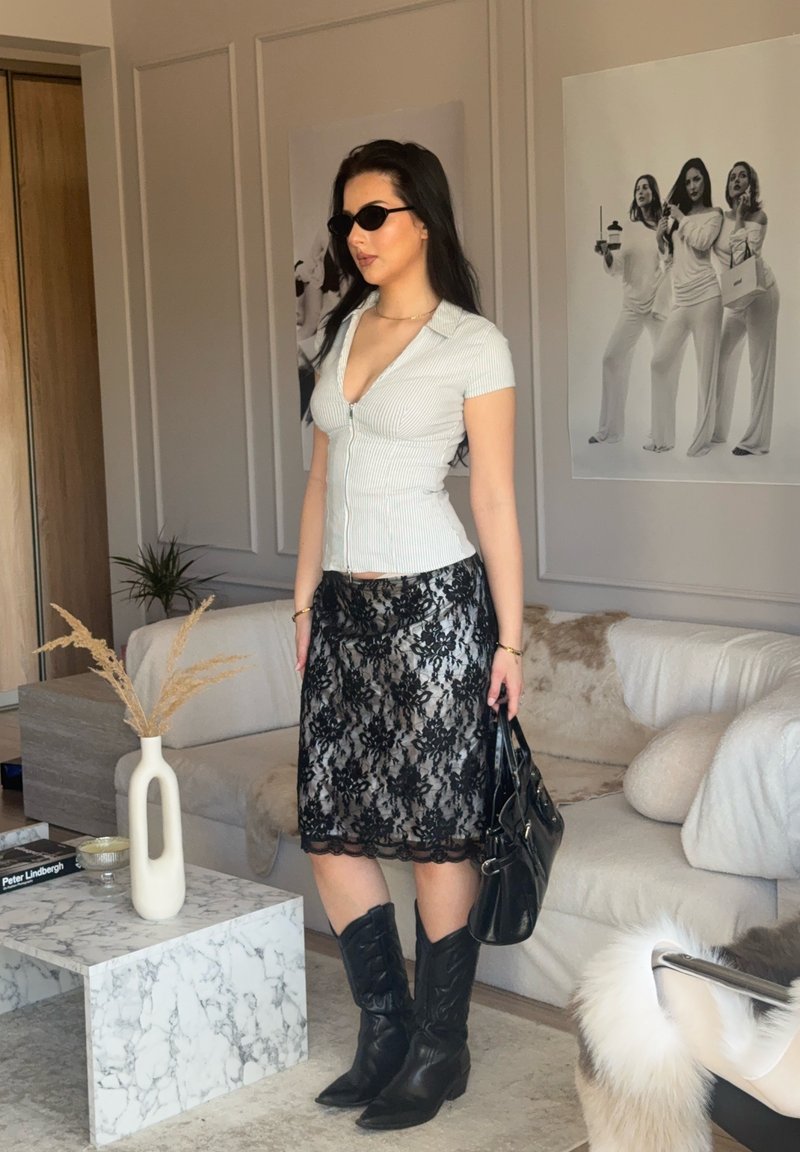 Femme portant des lunettes de soleil, un haut blanc ajusté, une jupe en dentelle noire et des bottes noires, tenant un sac à main noir dans un salon moderne.