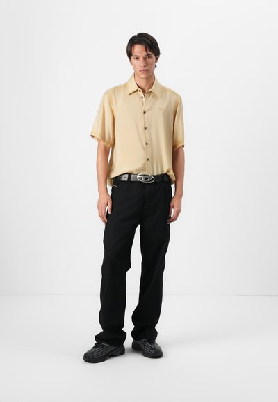 Camisa de manga corta, abotonada en un amarillo pálido, combinada con pantalones negros de corte holgado y zapatos negros. La camisa presenta un logo sutil.