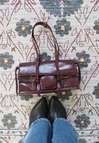 Sac à main en cuir verni bordeaux de forme rectangulaire, avec deux anses, une poche avant à rabat, posé sur un tapis à motifs.