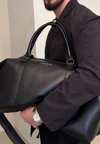Homme en blazer noir tenant un grand sac en cuir noir Valentino avec deux poignées et une montre argentée au poignet.