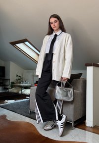Hvit strikket cardigan, skjorte med krage, svart slips, svarte bukser med hvite striper, sølvveske, hvite/grå joggesko og minimalistisch innredning.