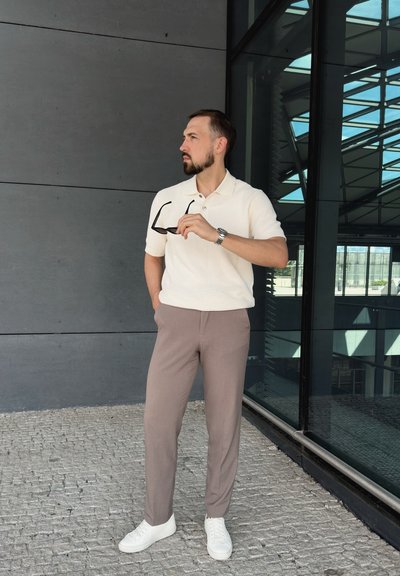 Camisa polo de textura beige, pantalones topo y zapatillas blancas. Sosteniendo gafas de sol. Fondo arquitectónico moderno con detalles de cristal y metal.