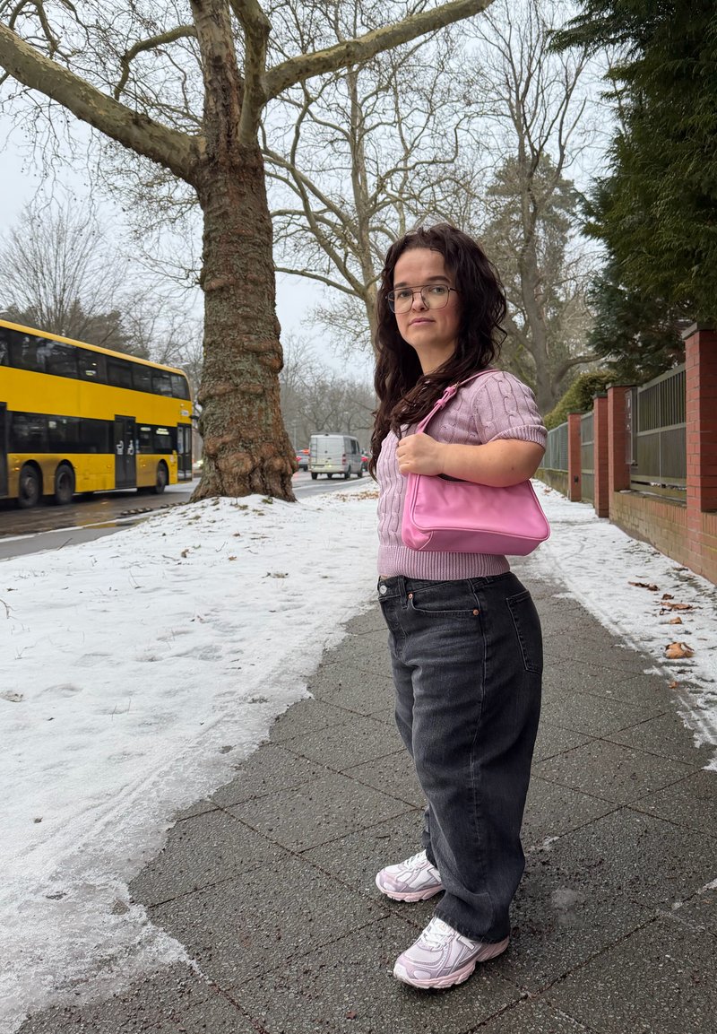 Femme portant des lunettes, un pull rose et un jean noir, tenant un sac à main rose, debout sur un trottoir enneigé près d'un bus à impériale jaune.