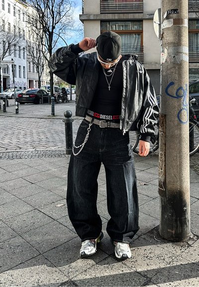 Chaqueta bomber de cuero negro con mangas a rayas, top negro ajustado, pantalones de mezclilla oscuros y holgados, accesorios con cadenas y un cinturón de marca. Zapatillas plateadas.
