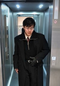 Jeune personne portant des lunettes, un manteau noir, des gants et une cravate, debout à l'intérieur d'un ascenseur moderne aux parois métalliques et aux éclairages au plafond.