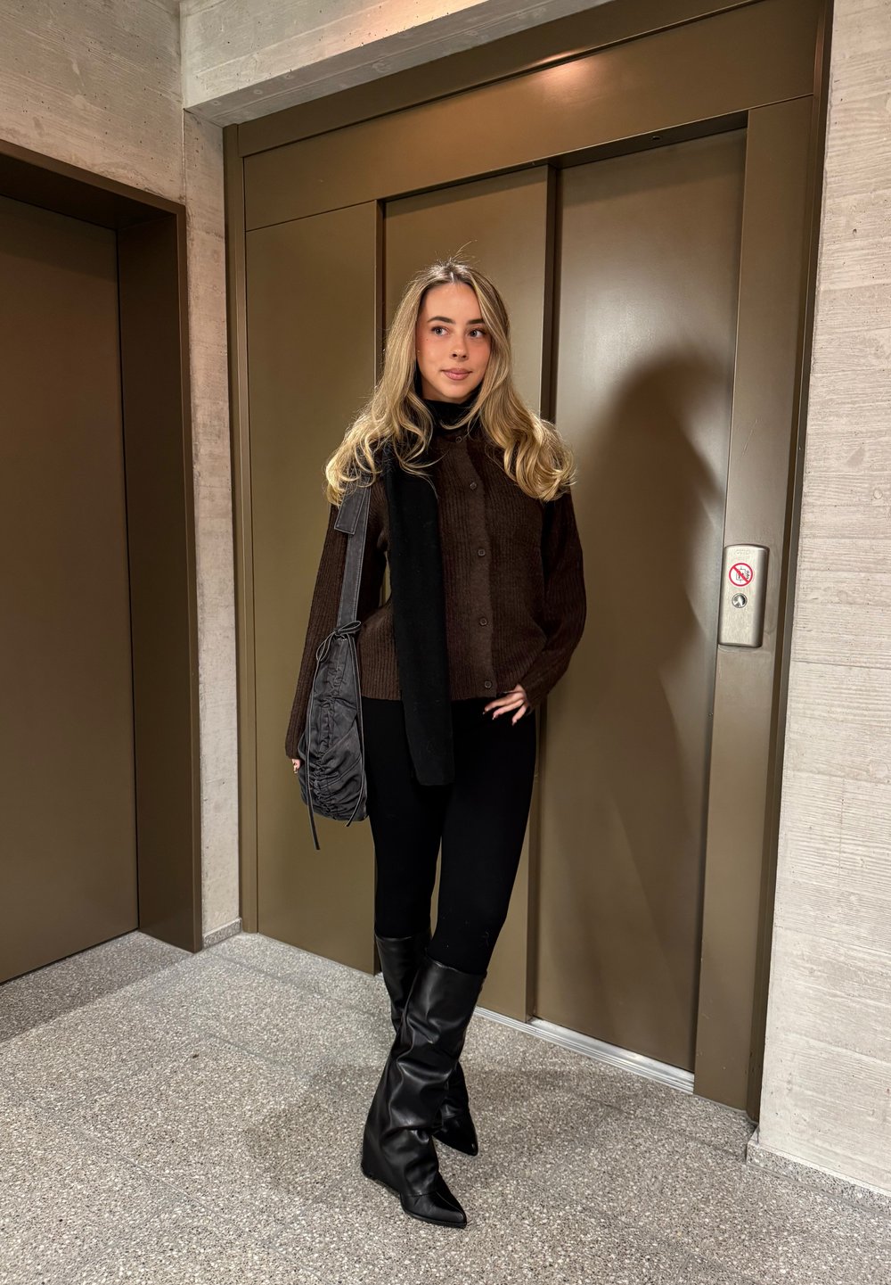 Barna bordázott gombos pulóver fekete sállal rétegezve, fekete leggingssel és térdig érő, hegyes orrú csizmával párosítva. Szürke táska, lift háttere.