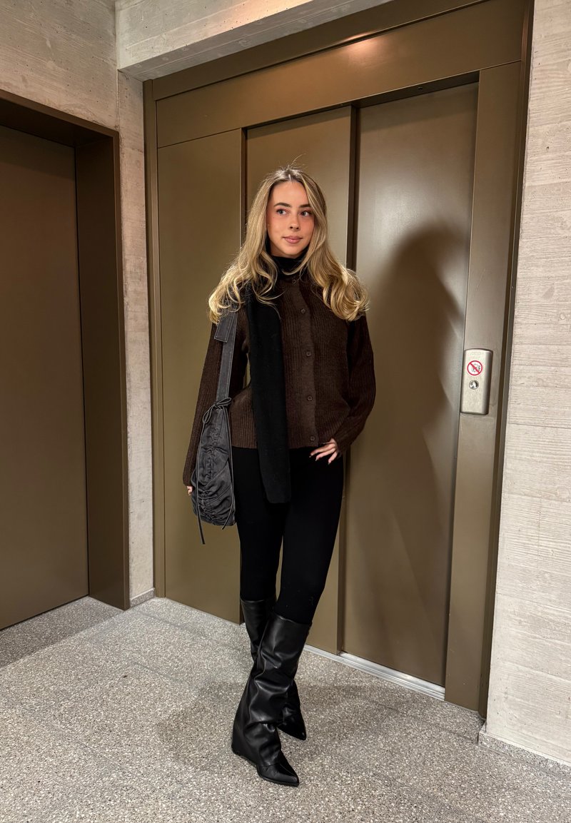 Brauner gerippter Strickpullover mit Knöpfen, darüber ein schwarzer Schal, kombiniert mit schwarzen Leggings und kniehohen Stiefeln mit spitzen Zehen. Graue Tasche, Hintergrund mit Aufzug.