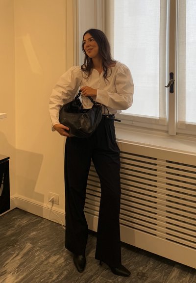 Mujer de pie en el interior junto a una ventana, vestida con una blusa blanca de mangas abullonadas, pantalones negros de cintura alta, botas negras y sujetando un bolso negro.