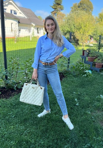 Bolso acolchado blanco con acentos dorados, jeans de mezclilla y una camisa de botones azul claro. El entorno es un área cubierta de hierba con plantas al fondo.