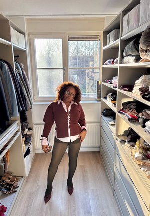 Mujer con cabello rizado y gafas posando en un vestidor con estantes organizados de ropa, zapatos y accesorios, iluminado por la luz natural de la ventana.