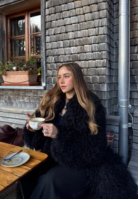 Jeune femme aux longs cheveux portant un manteau en fourrure noire, tenant une tasse de café, assise à une table en bois devant un bâtiment recouvert de bardeaux près d'une fenêtre.