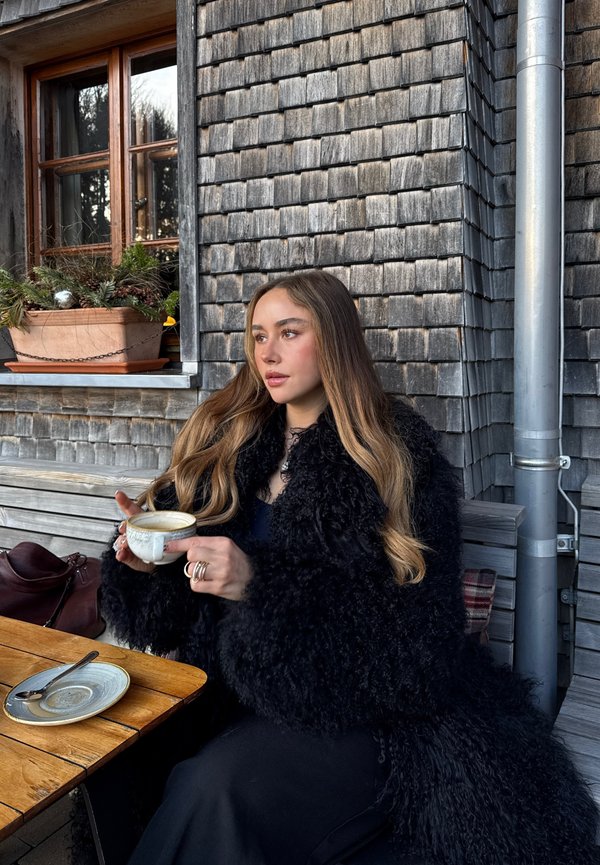 Junge Frau mit langen Haaren in einem schwarzen Pelzmantel, die eine Kaffeetasse hält und an einem Holztisch vor einem mit Schindeln verkleideten Gebäude in der Nähe eines Fensters sitzt.