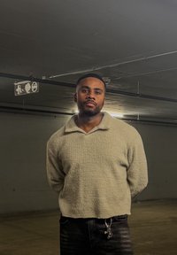 Homme portant un pull beige texturé et un pantalon noir, debout les mains derrière le dos dans un parking souterrain faiblement éclairé.