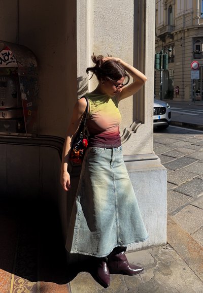 Mujer joven con el cabello recogido, gafas de sol, top sin mangas tie-dye, falda vaquera y botas moradas, de pie junto a una columna de piedra bajo la luz del sol.