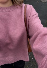 Jasnoróżowy dziergany sweter z prążkowanym okrągłym dekoltem i długimi rękawami, zestawiony z brązową torbą na ramię, na tle ciemnej, teksturowanej nawierzchni.