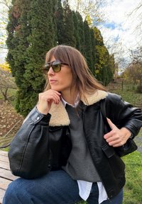 Crna kožna jakna s bež ovratnikom od ovčje kože, siva majica ispod, bijela košulja, tamne traperice i sunčane naočale u obliku kornjačevine.