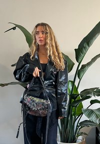 Femme aux longs cheveux bouclés portant une veste noire brillante oversize et un pantalon noir, tenant un sac à bandoulière coloré à motifs de texte à l'intérieur, à côté de plantes.