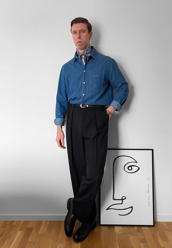 Άνδρας με μπλε πουκάμισο κουμπωτό και μαύρο παντελόνι στέκεται με το χέρι στην τσέπη δίπλα σε κορνιζαρισμένη αφηρημένη τέχνη προσώπου πάνω σε ξύλινο πάτωμα.