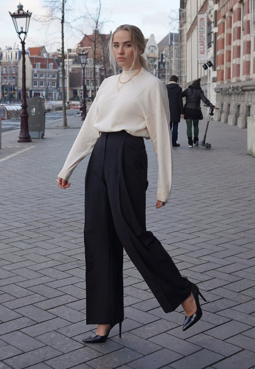 Suéter crema, pantalones negros de cintura alta y tacones de aguja negros. Modelo en un entorno urbano con pavimento estampado y edificios al fondo.