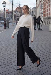 Suéter crema, pantalones negros de cintura alta y tacones de aguja negros. Modelo en un entorno urbano con pavimento estampado y edificios al fondo.