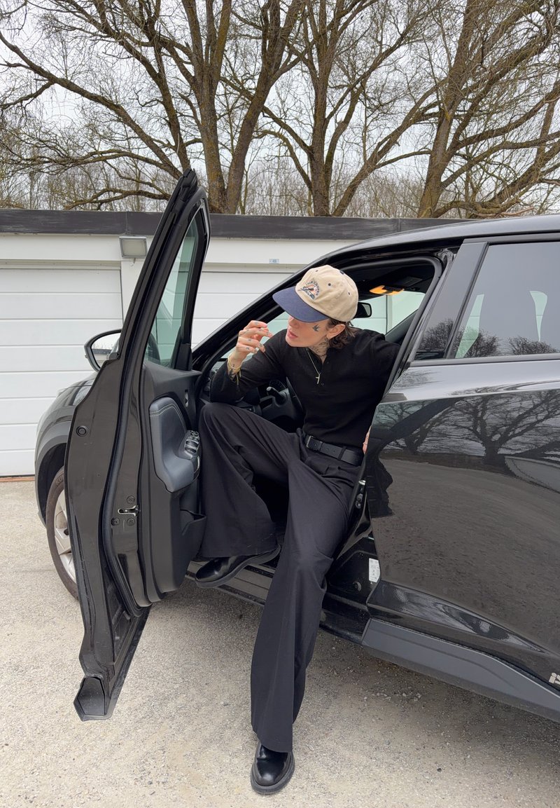 Personne portant une casquette beige et une tenue noire assise de côté dans une voiture noire avec la porte du conducteur ouverte en extérieur.