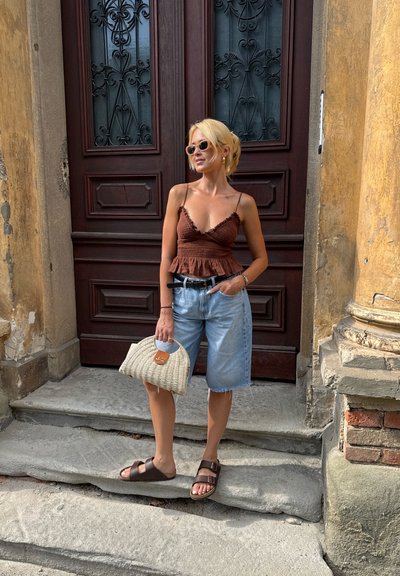 Top marrón sin mangas con dobladillo acampanado, pantalones cortos de mezclilla azul claro, sandalias marrones y un bolso de mimbre. De pie en unos escalones de piedra cerca de una puerta.