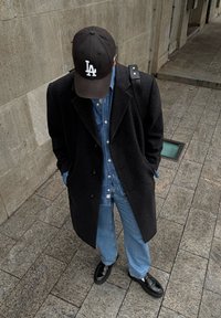 Μαύρο μάλλινο παλτό, μπλε τζιν πουκάμισο και παντελόνι, μαύρα δερμάτινα παπούτσια και μαύρο καπέλο μπέιζμπολ με λευκή κεντημένη λεπτομέρεια, που στέκεται σε πέτρινο πλακόστρωτο.