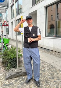 Črn najlonast telovnik z več žepi, bela majica, ohlapne kavbojke in črne škornje. V ozadju so zelenje in urbane oznake.