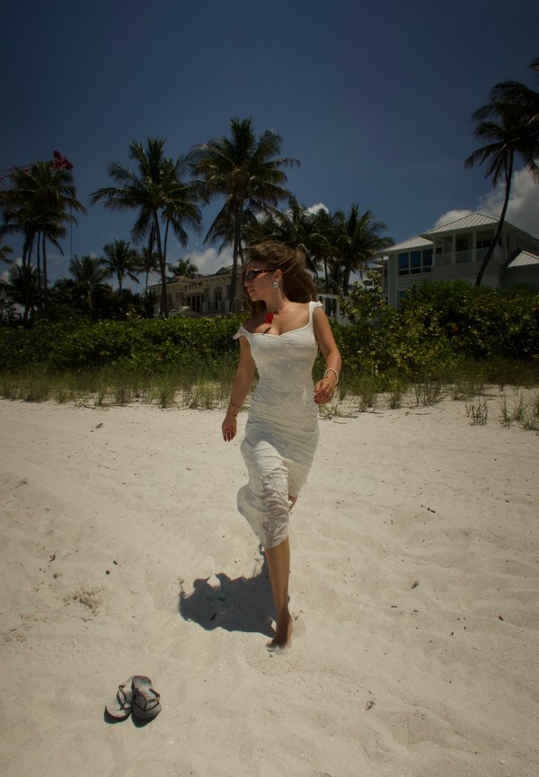 Frau in weißem Kleid geht barfuß am Sandstrand nahe Palmen und Strandhäusern unter klarem Himmel.
