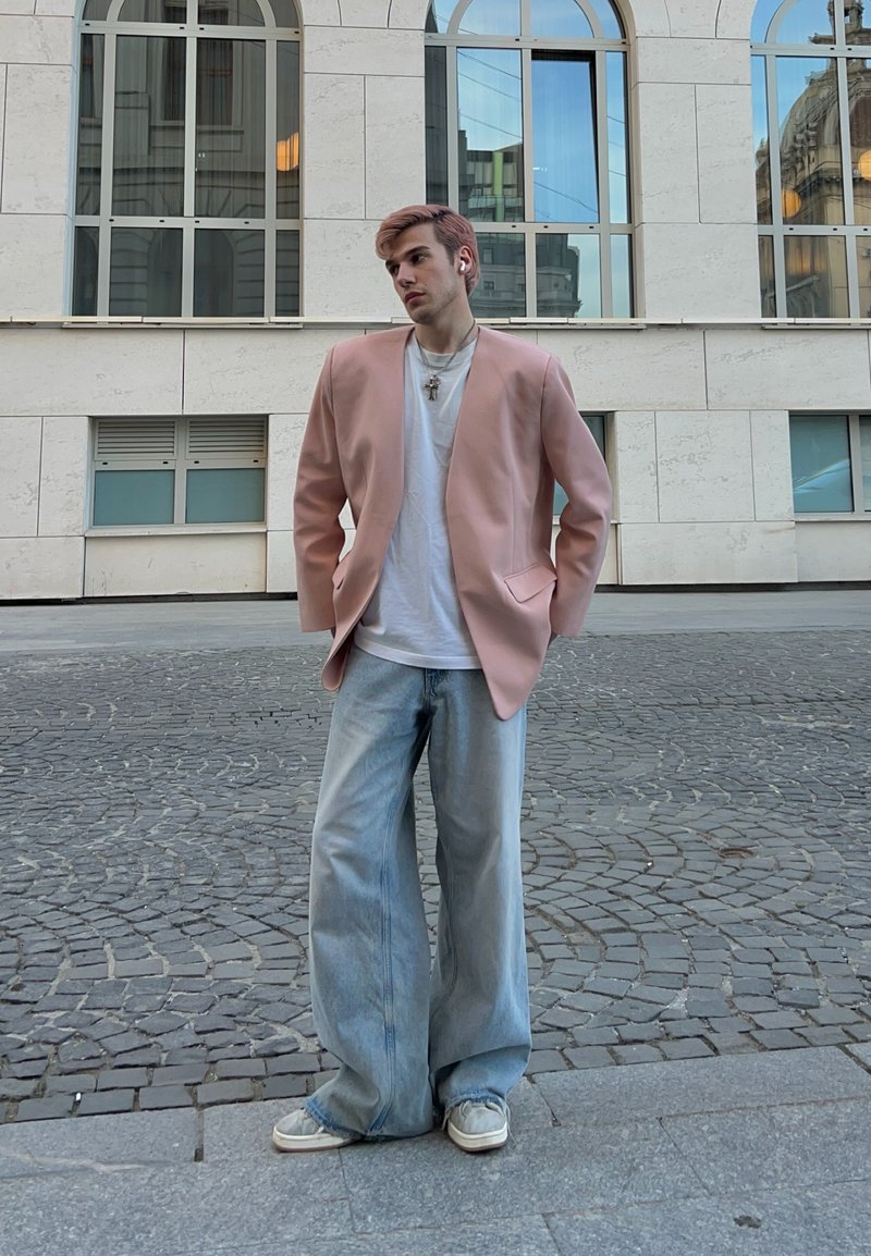 Jeune personne debout dans une rue pavée, portant un blazer rose oversize, une chemise blanche, un jean large, des baskets et des écouteurs sans fil.