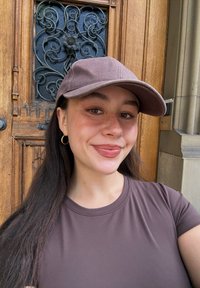 Brun baseballkasket, brun top med korte ærmer, langt mørkt hår, fregner. Trædør med indviklet metalarbejde i baggrunden.