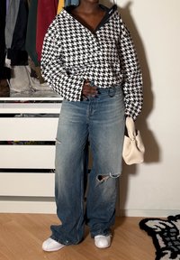 Personne portant une veste pied-de-poule noir et blanc, un jean large déchiré, des chaussures blanches et tenant un sac à main beige à l'intérieur.