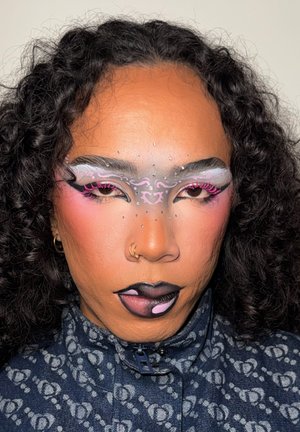Persona con cabello rizado negro, maquillada de manera elaborada en tonos rosa, blanco y gris en el rostro, con arte labial artístico en negro y rosa, vestida con ropa oscura con patrones.