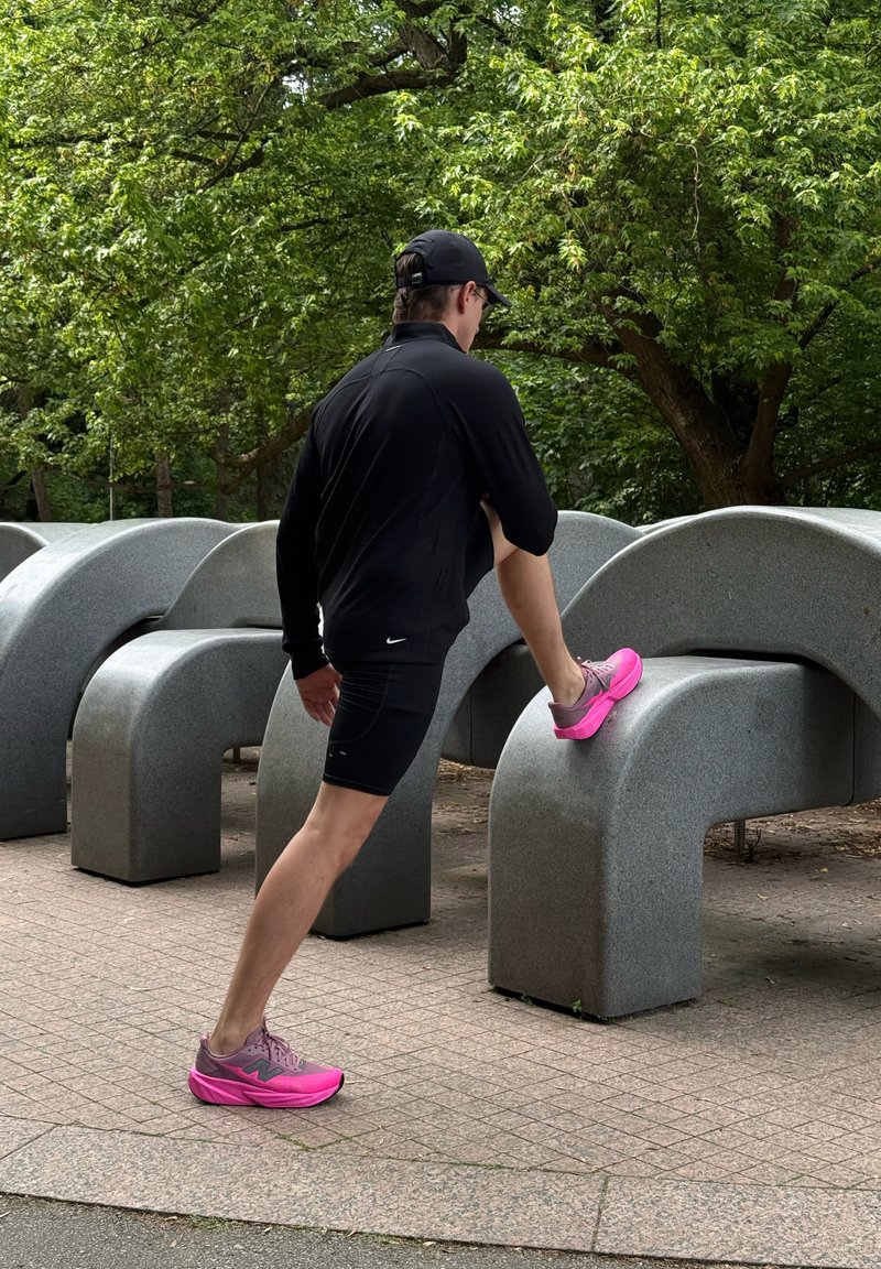 Homme en tenue sportive noire et chaussures roses, étirant une jambe sur une structure en béton gris incurvée à l'extérieur avec des arbres verts en arrière-plan.